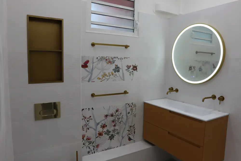 Salle de bain rénovée en Guadeloupe : niche dorée, carrelage fleuri, miroir rond et meuble vasque suspendu